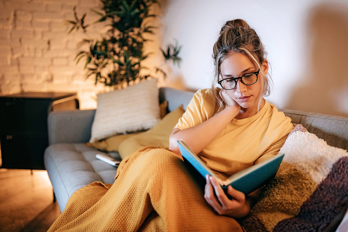 A woman curling up on her couch to read a book to relieve stress