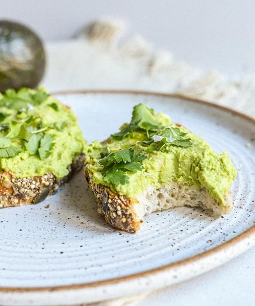 two slices of avocado toast laying on a plate with one having a bite taken out of it. 