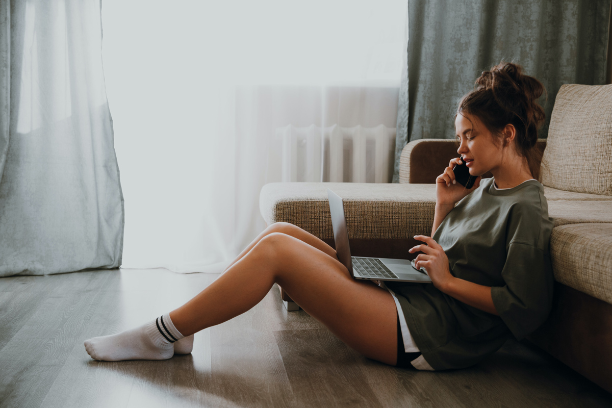 A girl sitting by her couch talking on the phone and her laptop looking stressed