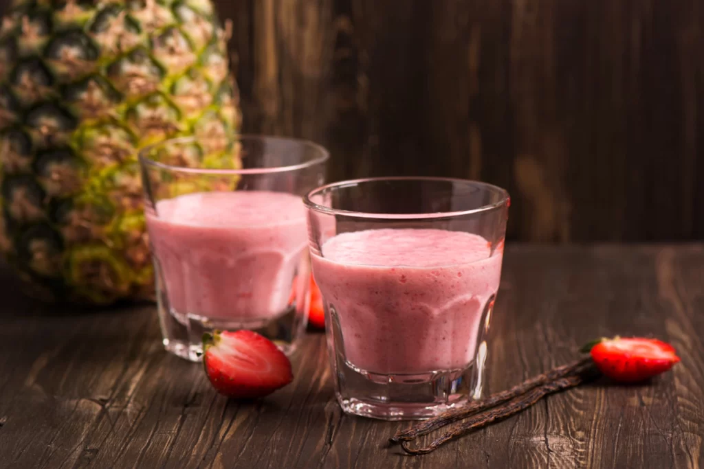 Two glasses of a strawberry pineapple smoothie on a set with a decorative pineapple and loose strawberries on the table